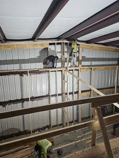 Construction workers inside a building frame, installing framing and working with power tools; steel beams overhead.