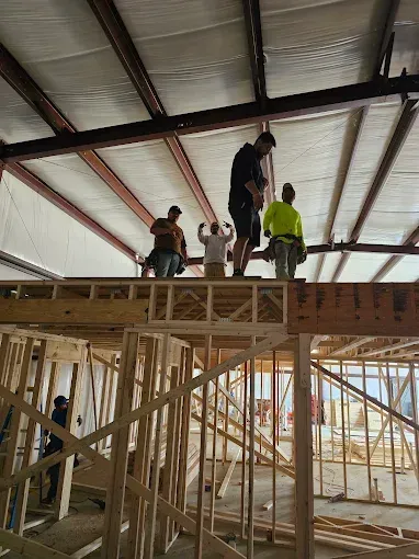 Construction workers framing a building; some on a wooden platform. Sunlight streams in.