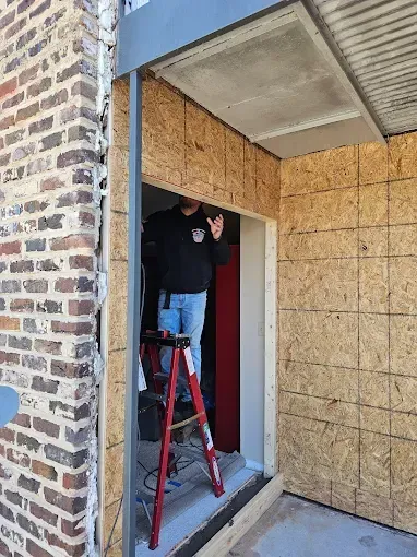 Man on ladder in doorway installing OSB paneling, brick building exterior.