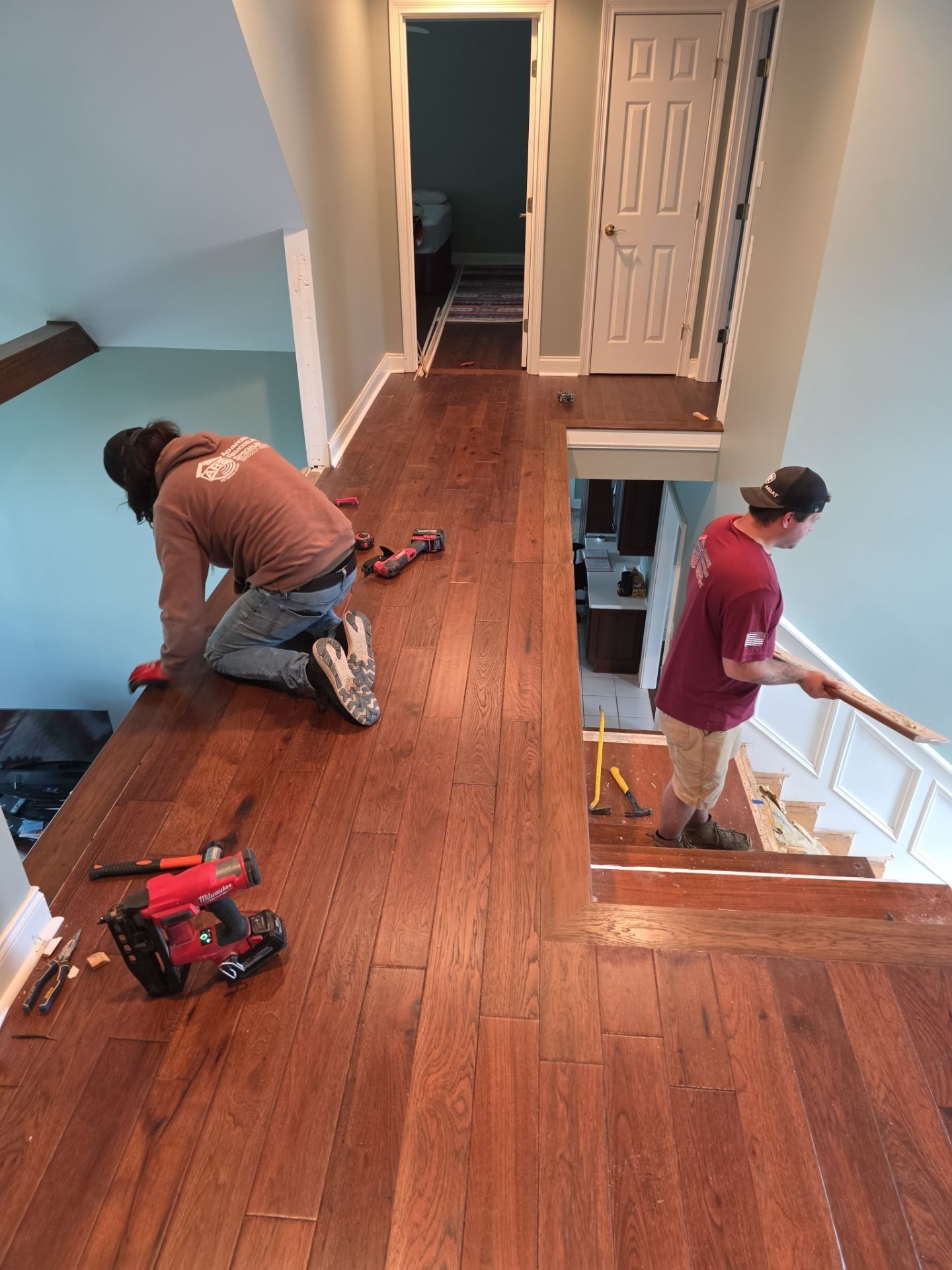 Two workers installing hardwood floors on a hallway and stairs, brown wood, light blue and white walls.