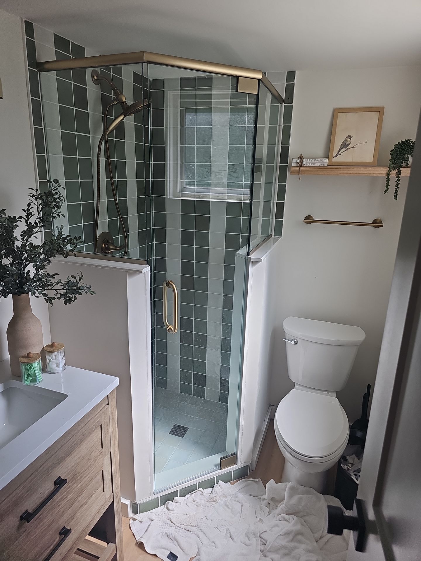 Green tiled shower with gold accents, next to a toilet, small wooden shelf with decor.