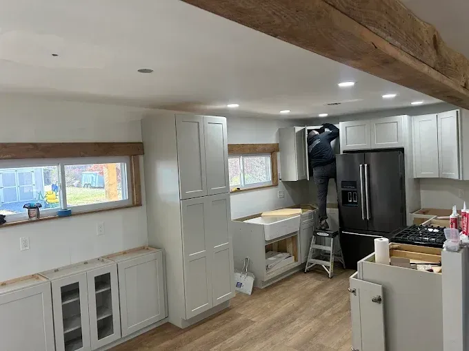 Kitchen renovation: a person on a step stool installs a cabinet. White cabinets, black refrigerator, wooden beams.