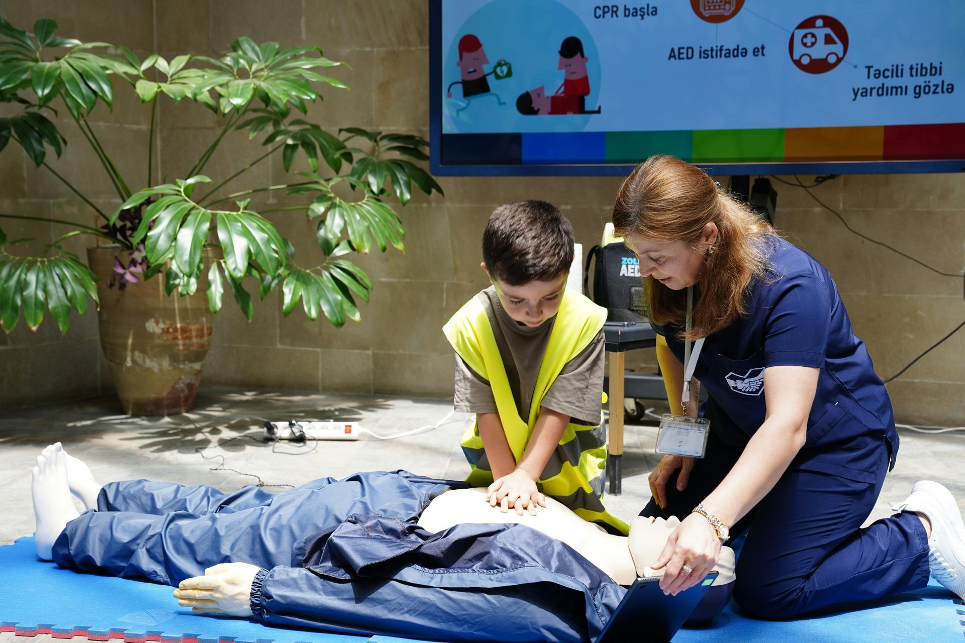 Woman instructs a child in CPR on a dummy. They are indoors, a presentation screen is visible.