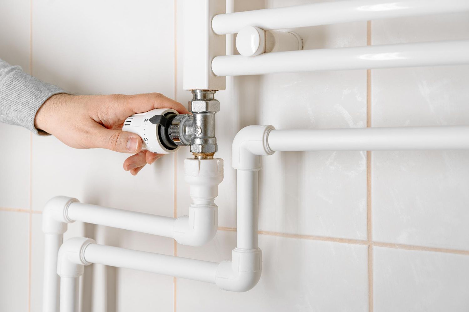 Hand turning a valve on white bathroom pipes against a tiled wall