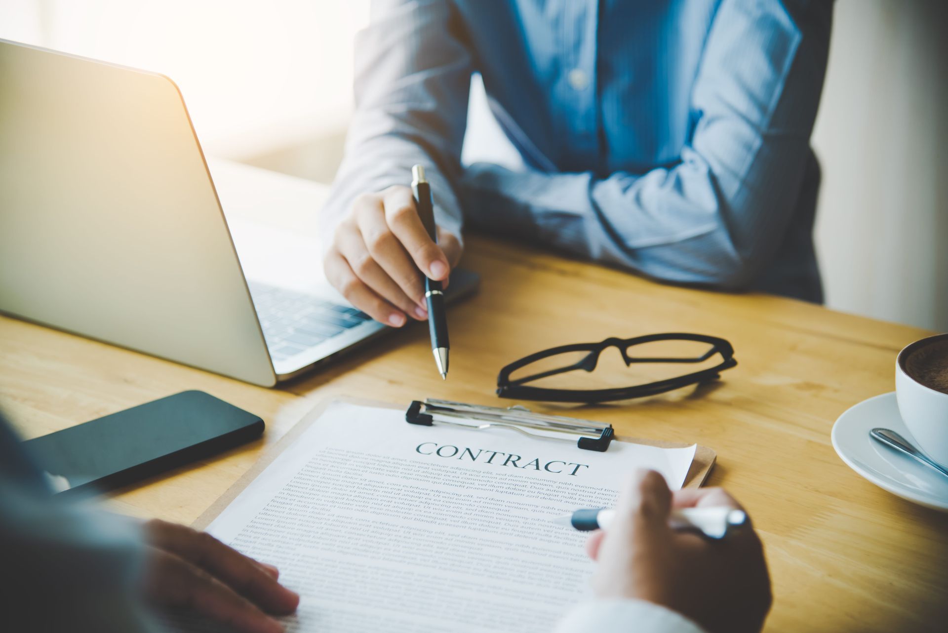 Person points at a contract on a desk with a laptop, glasses, and coffee cup nearby.