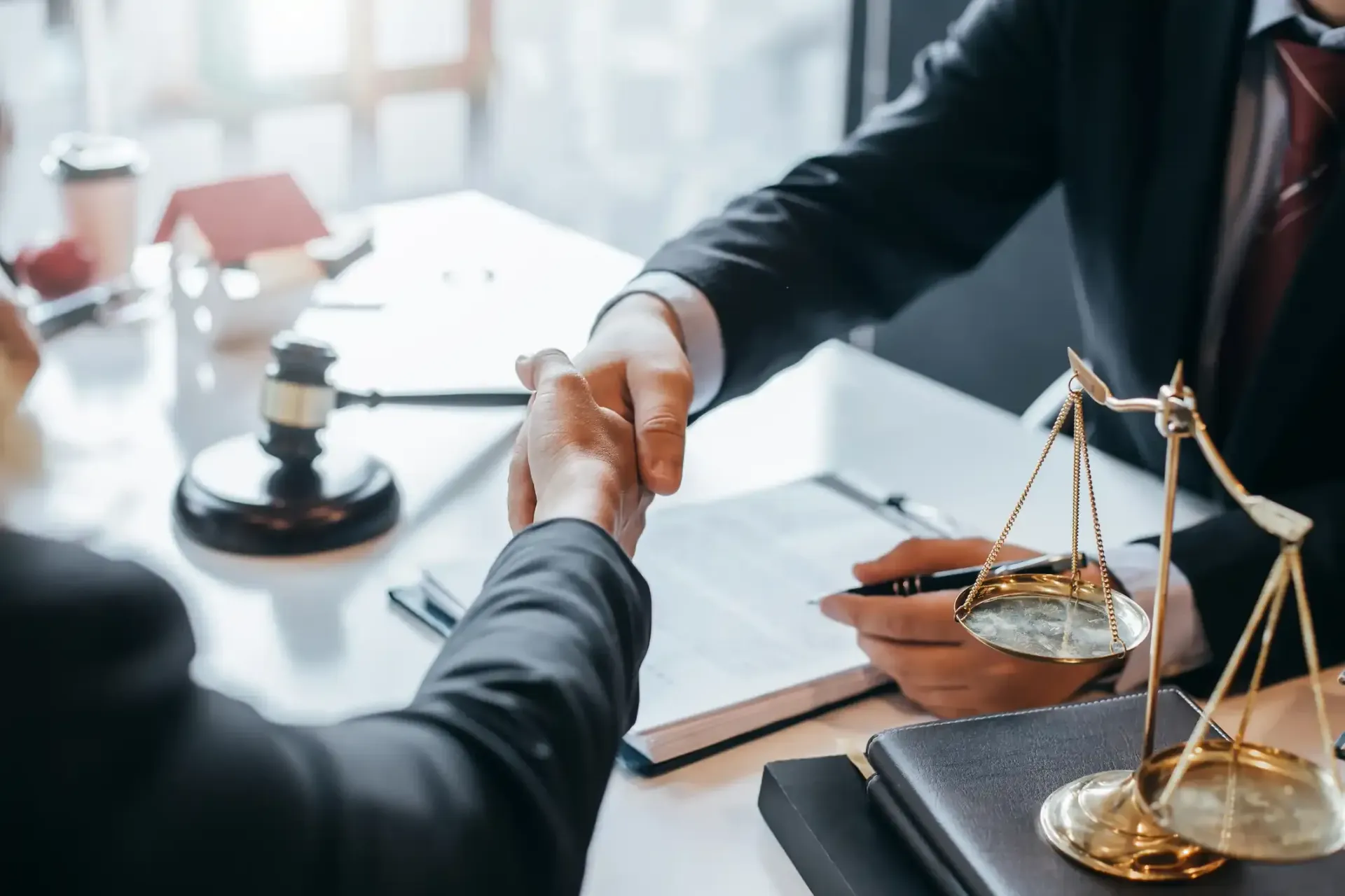 Two people in suits shaking hands at a desk with legal scales, documents, and a gavel.