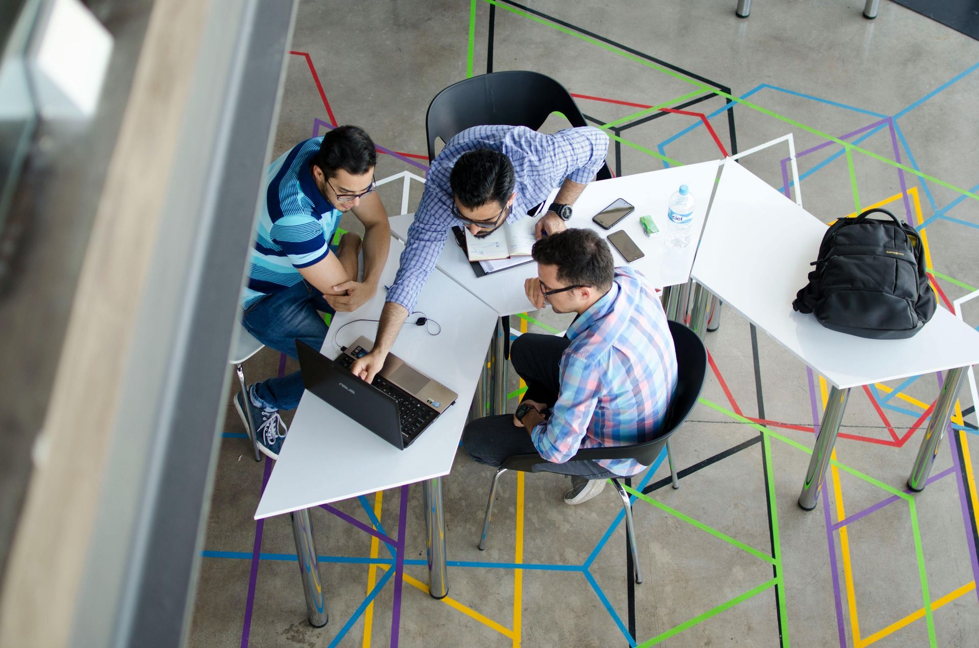 Three men collaborating around a laptop on a white table in an office. Colorful geometric floor design.