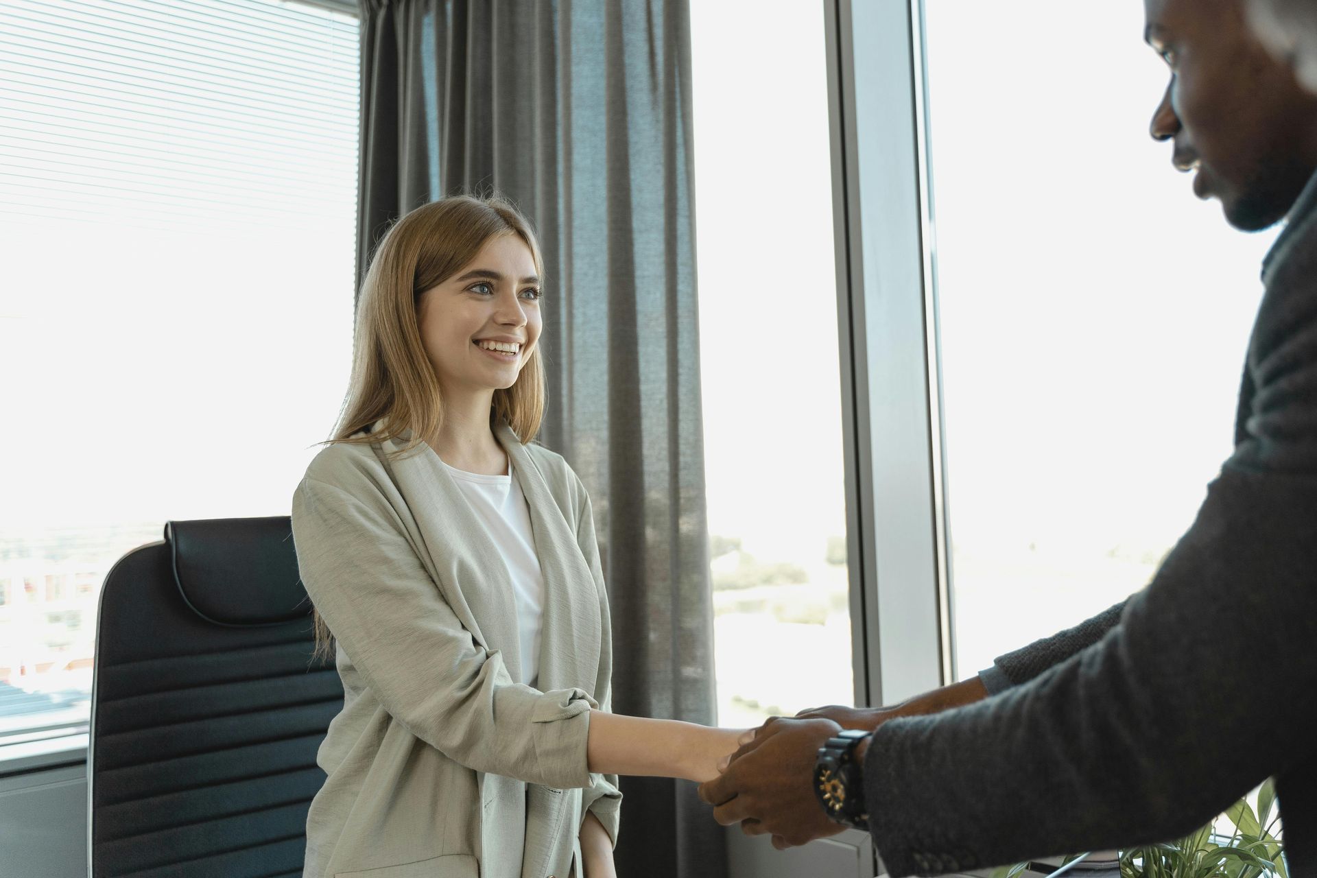 Woman in beige jacket shakes hands with a person in a suit, near a window in an office.