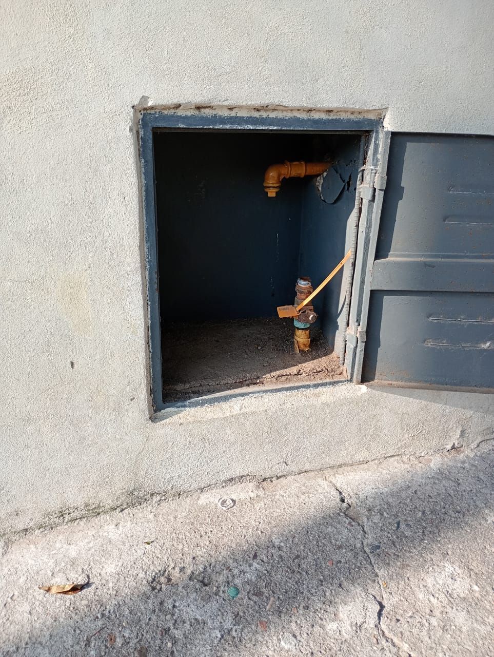 Abertura de pared rectangular de color gris con instalación de plomería en su interior; puerta metálica abierta.
