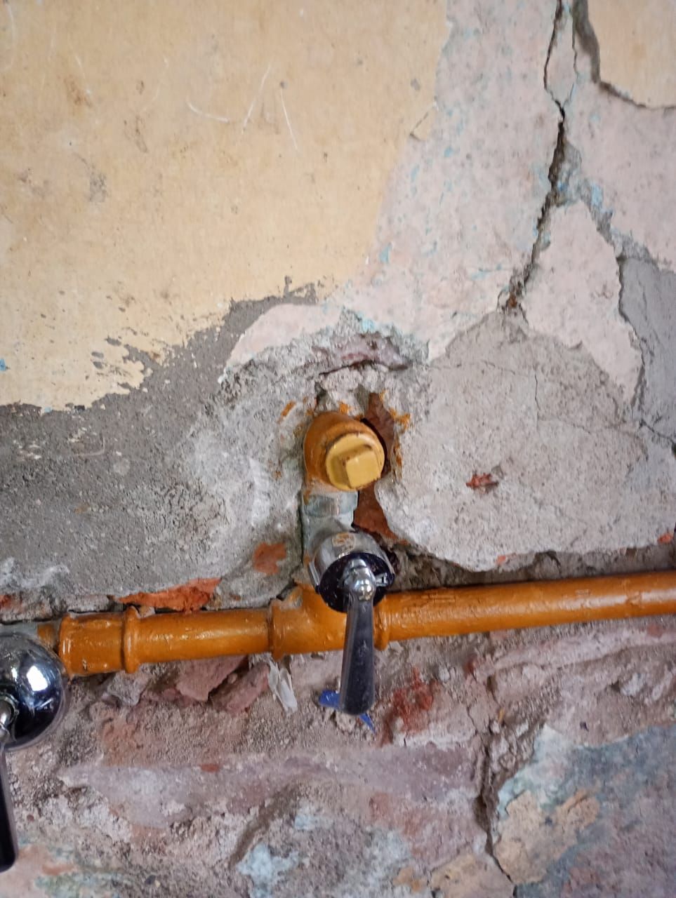 Tuberías de plomería y grifo de color naranja contra una pared dañada y texturizada.