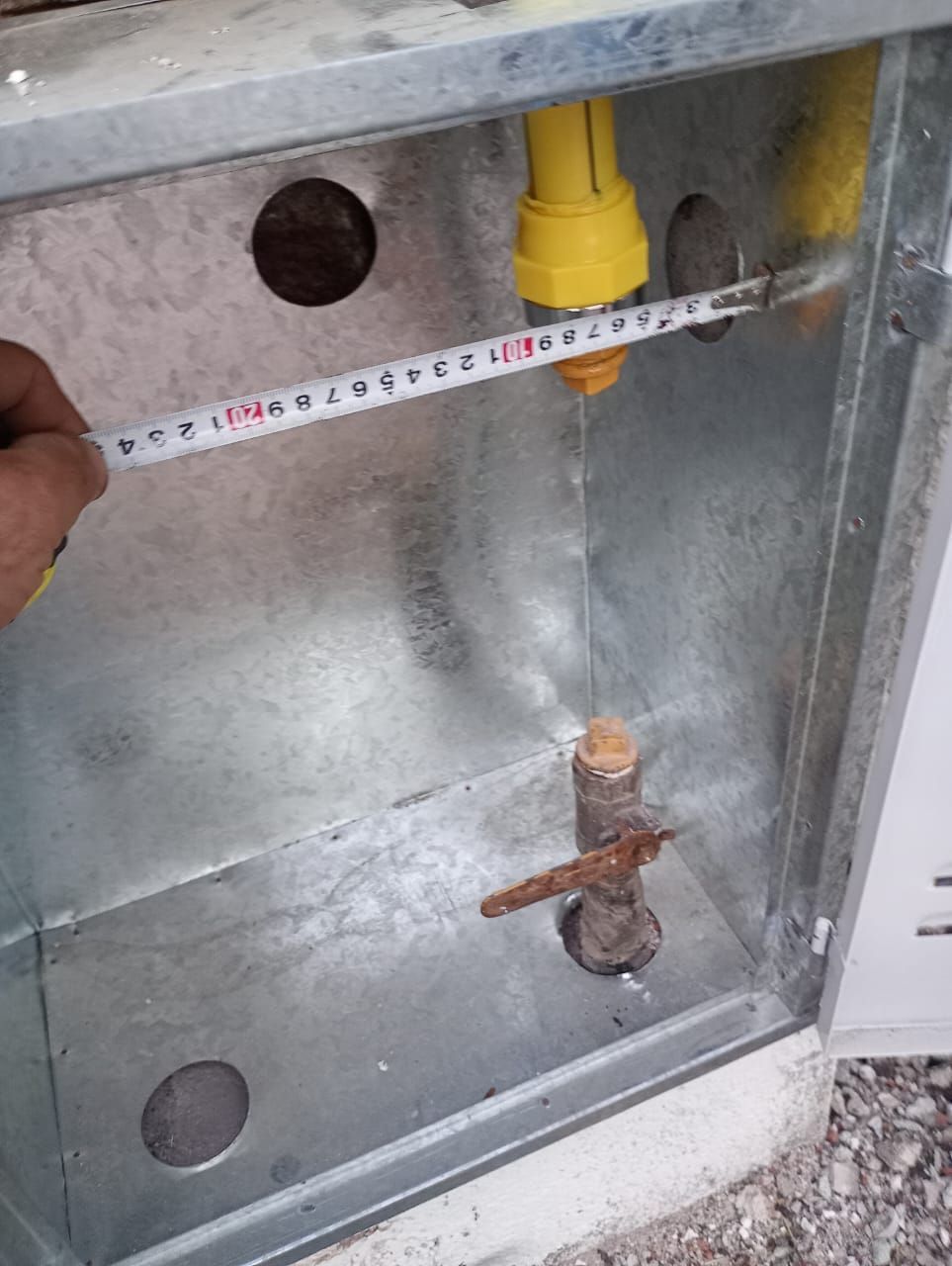 Una persona mide el interior de una caja metálica con una cinta métrica. Hay dos tapones amarillos y una tubería oxidada.
