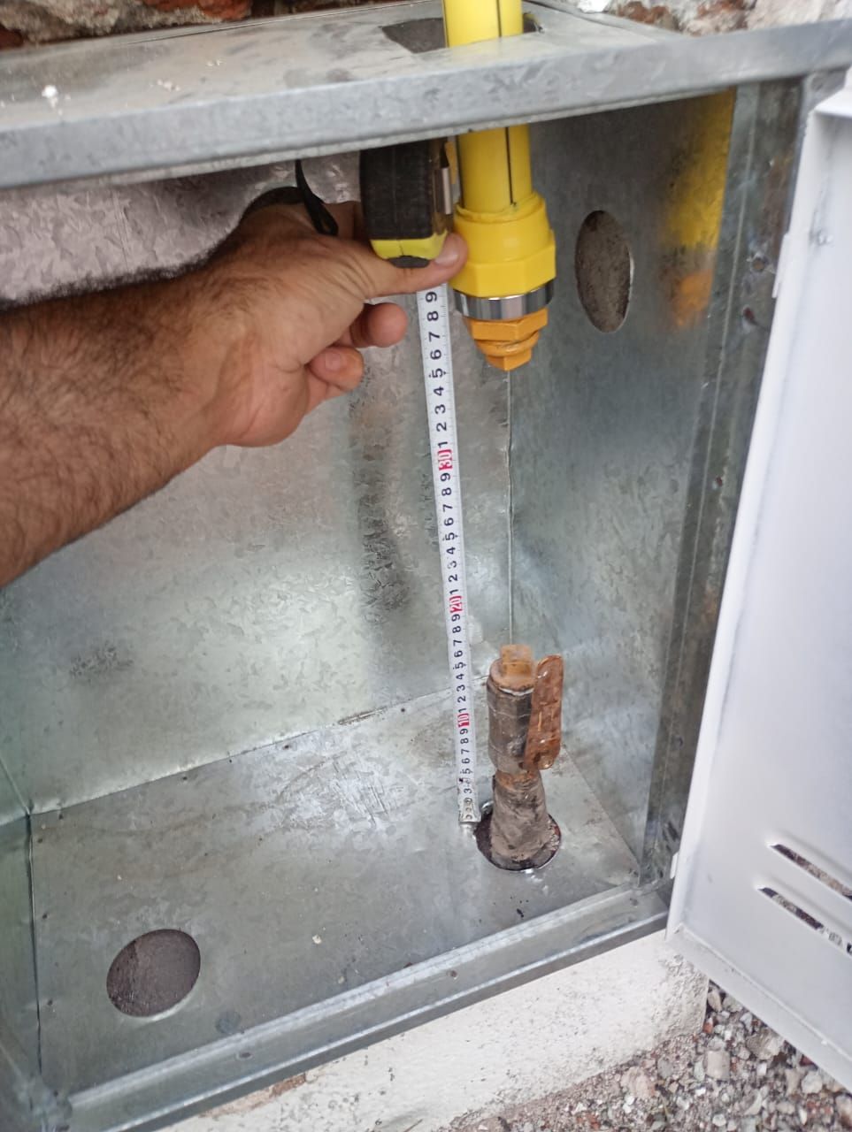 Persona midiendo una línea de gas dentro de una caja metálica con una cinta métrica. Se ven tuberías amarillas y oxidadas.