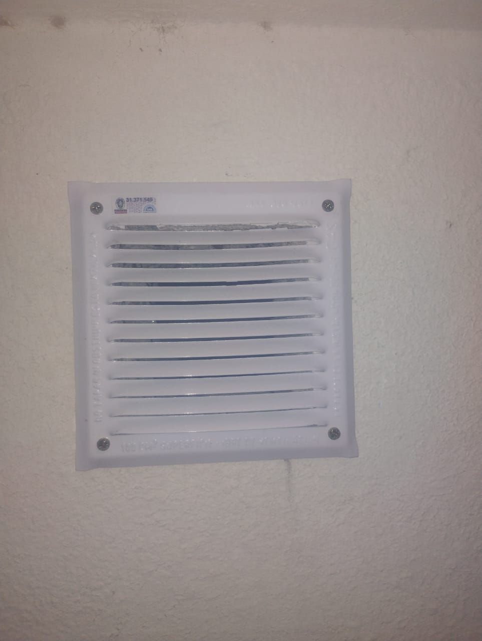 Cubierta de ventilación rectangular blanca con lamas horizontales sobre una pared blanca.