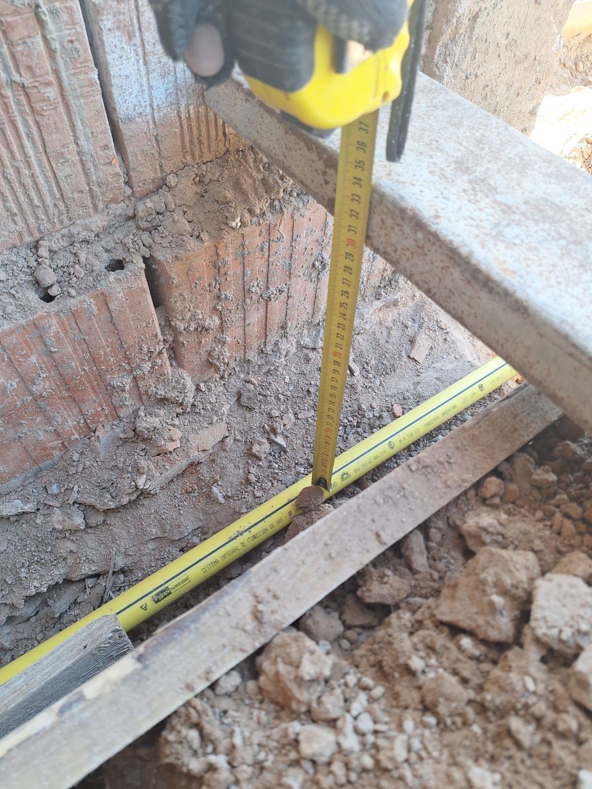 Una persona mide la profundidad en una zanja con una cinta métrica amarilla, junto a ladrillos y madera.