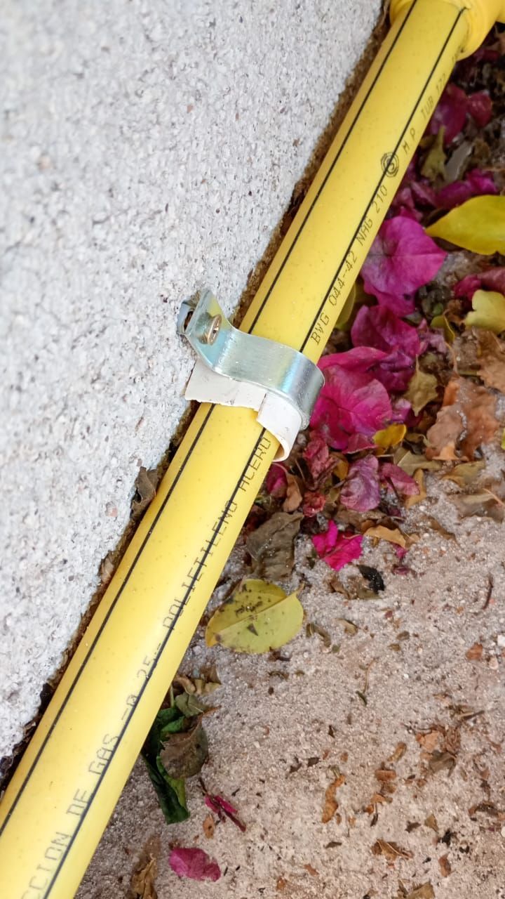 Tubo de gas amarillo fijado a una pared blanca con una abrazadera de metal, escombros en el suelo.