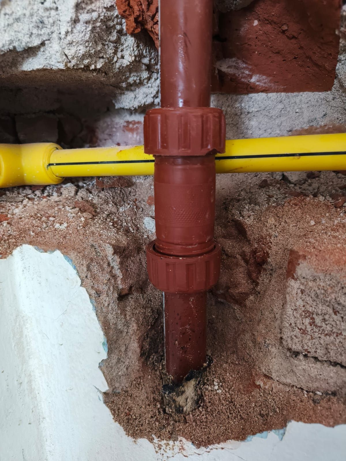 Tubo de plástico marrón con acoplamiento contra una pared de ladrillos, tubería de gas amarilla en el fondo.