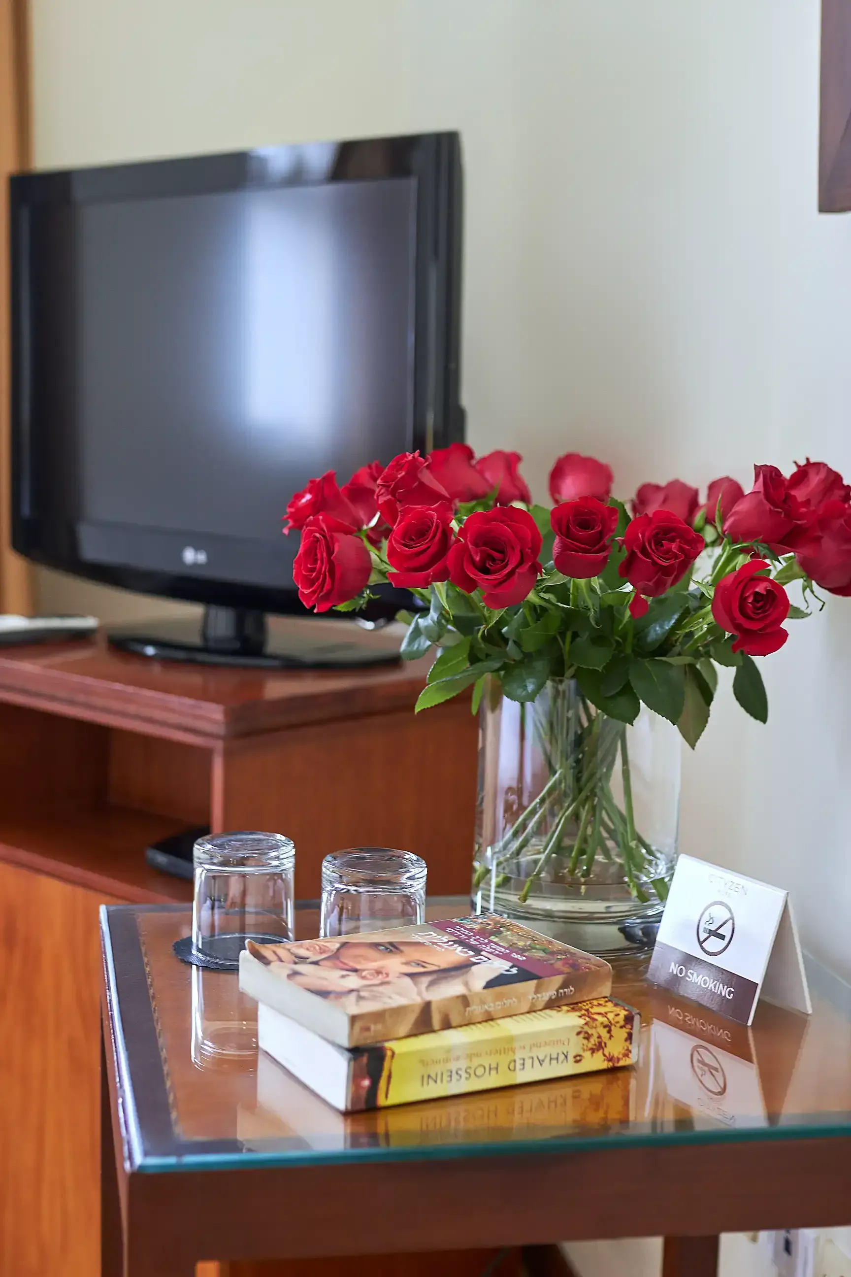 A vase of red roses sits on a glass table next to a television.