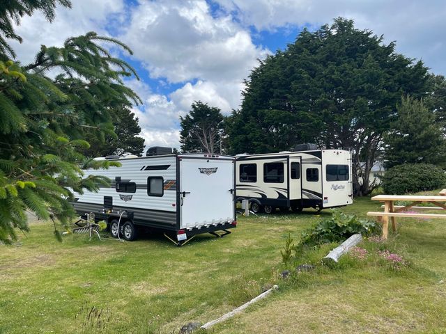 Two travel trailers parked in a grassy campsite surrounded by trees under a cloudy sky with a wooden picnic table nearby.
