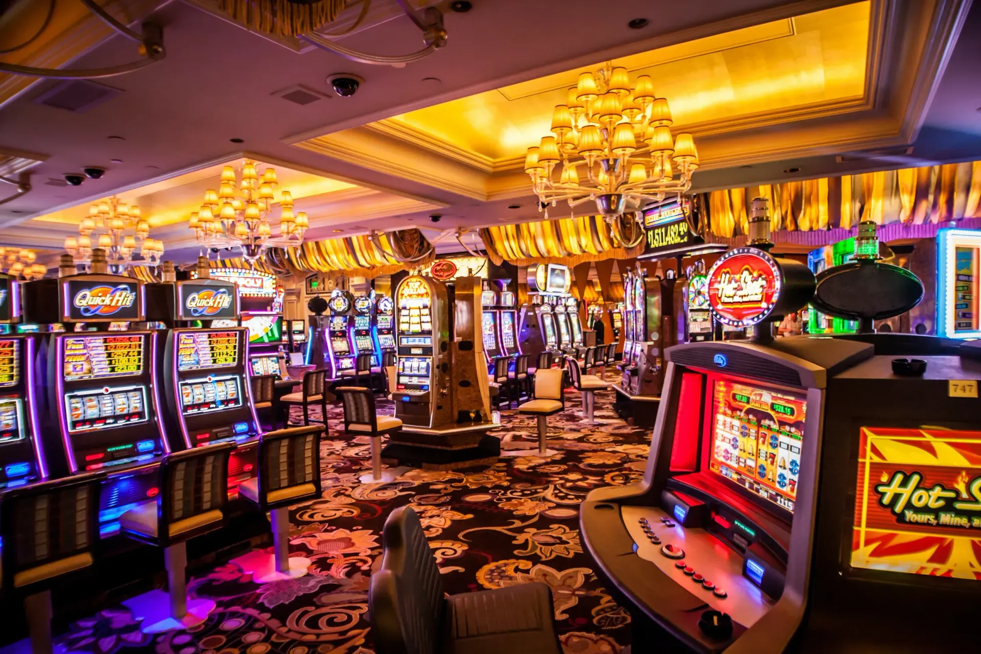Rows of lit slot machines in a dimly lit, carpeted casino floor featuring ornate, golden chandeliers.