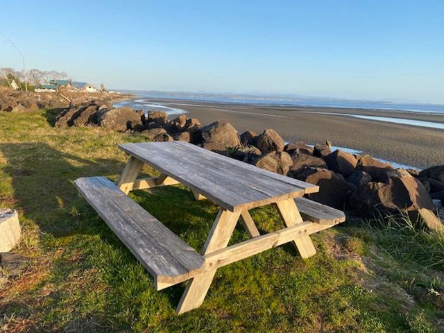 A weathered wooden picnic table sits on a grassy bank overlooking a rocky shoreline and the calm ocean at low tide.
