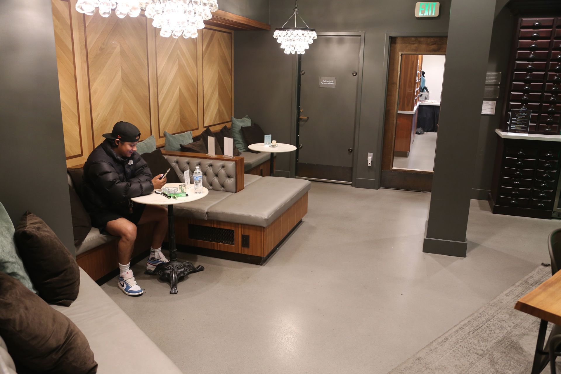 A man in a black jacket sits at a small table in a dimly lit lounge, looking at his phone.