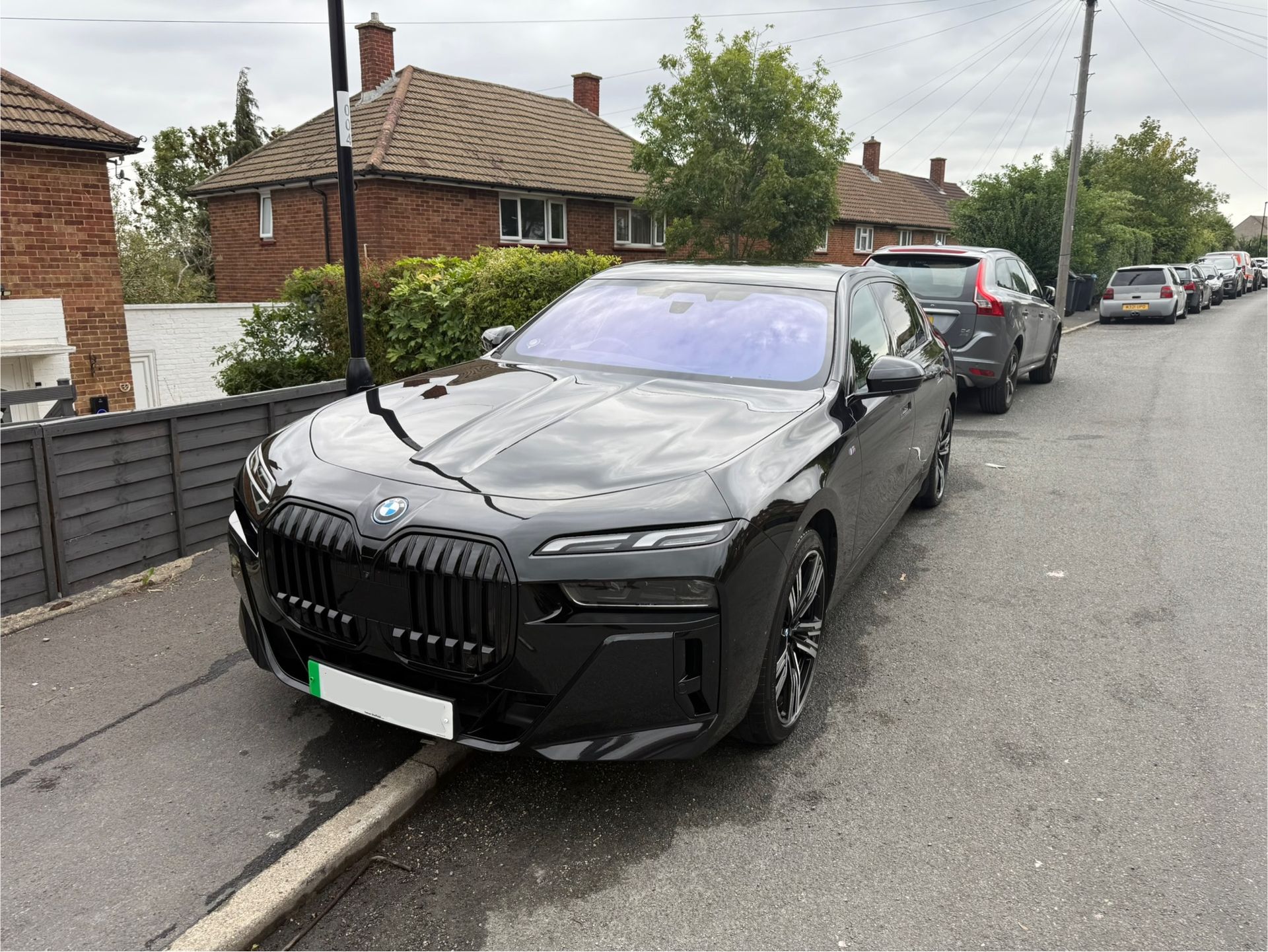 Black BMW sedan parked on a residential street; gray sky, parked cars, brick houses in background.