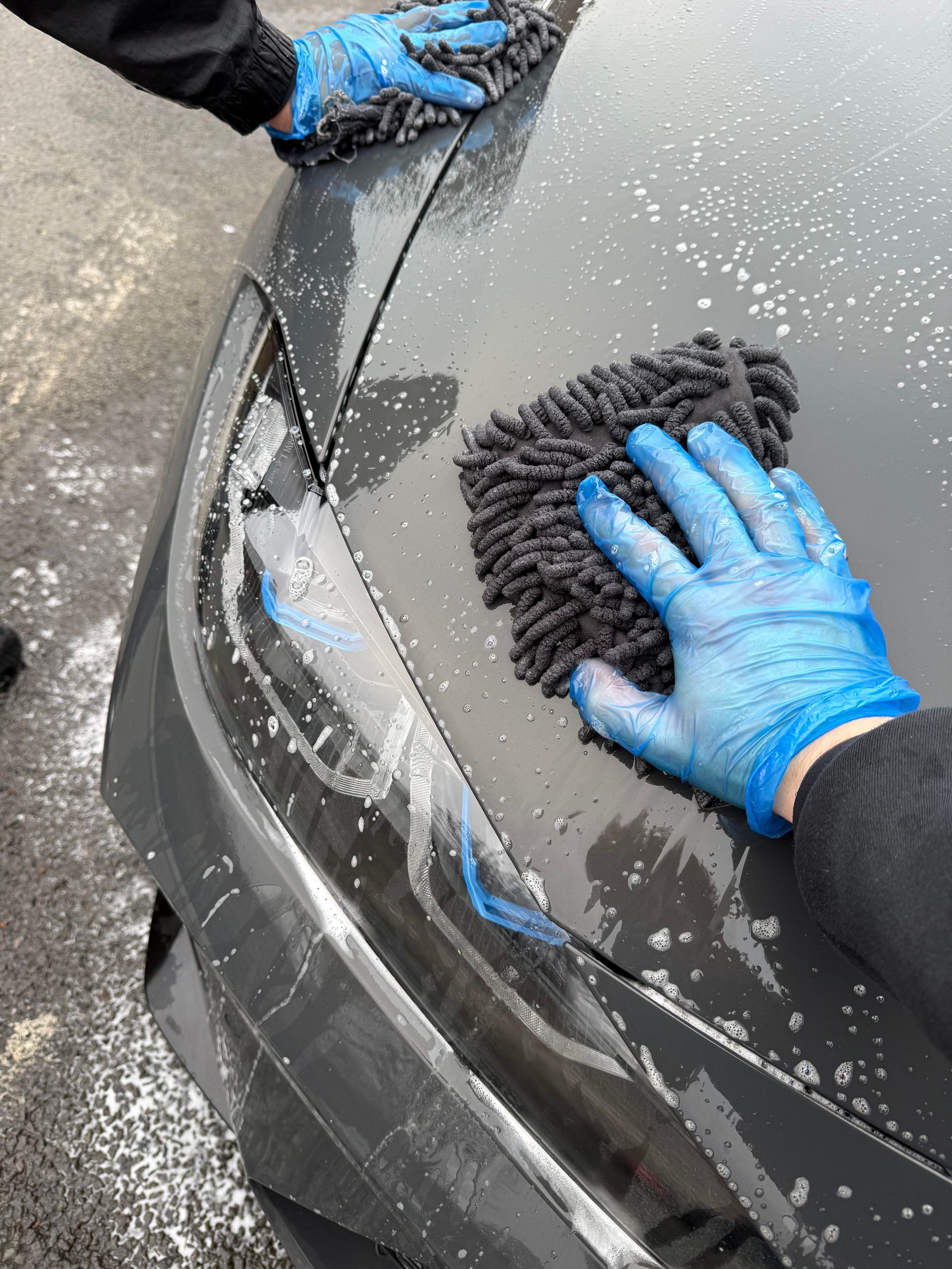 Hands in blue gloves washing a dark grey car with a fluffy microfiber mitt.