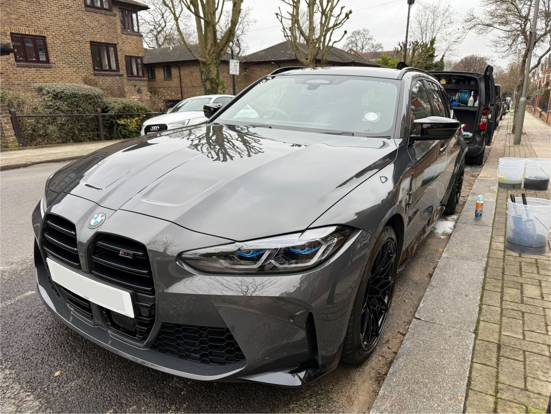 Gray BMW M3 parked on a street next to a sidewalk with other vehicles and buildings in the background.