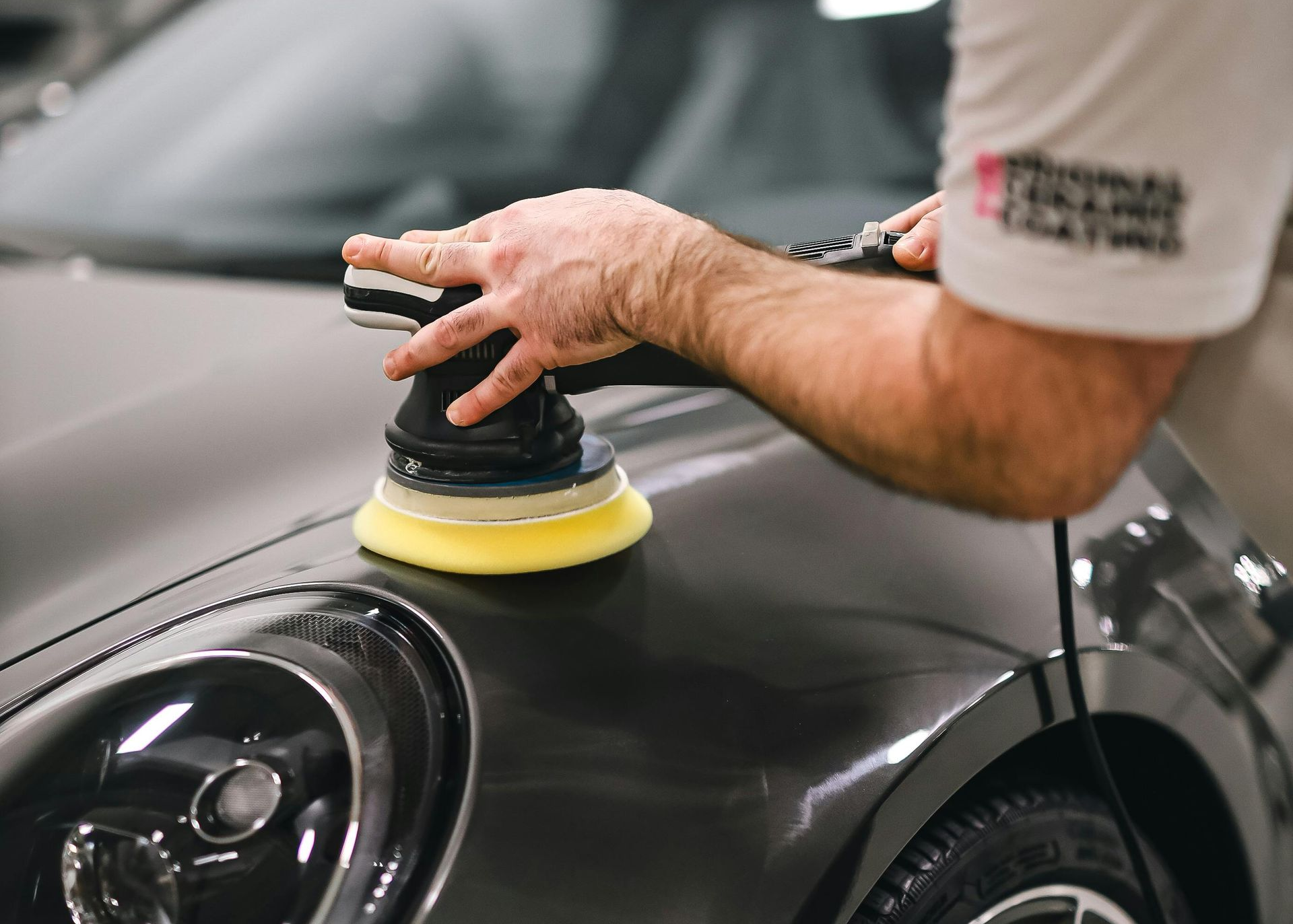 Person polishing dark gray car with a buffer, yellow pad attached.