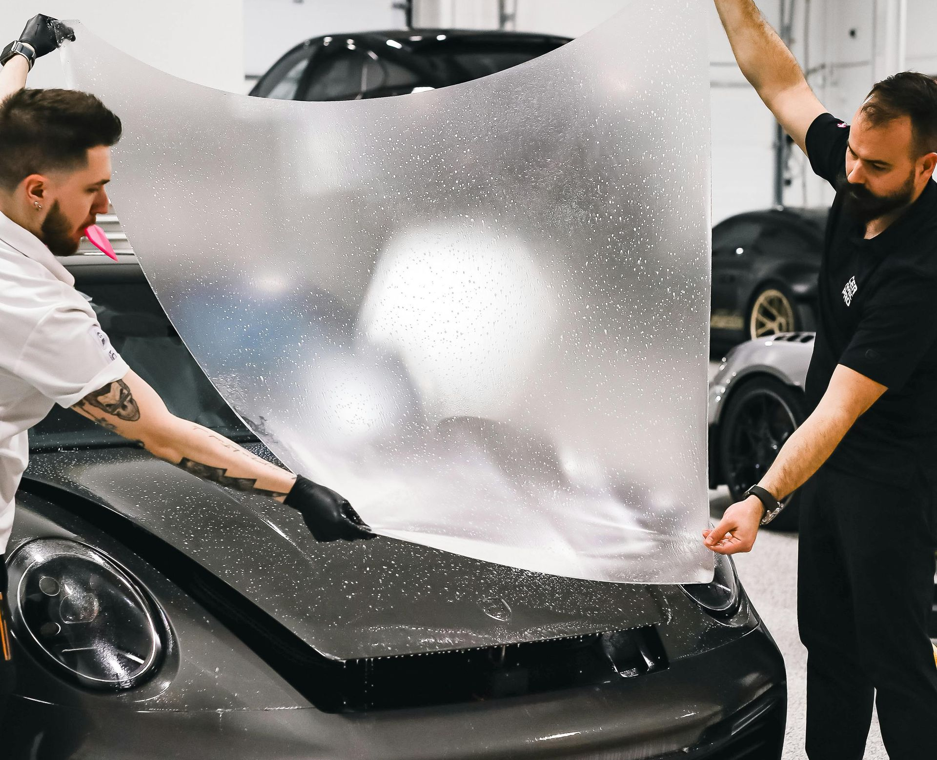 Two people applying clear protective film to a gray car hood in a shop.