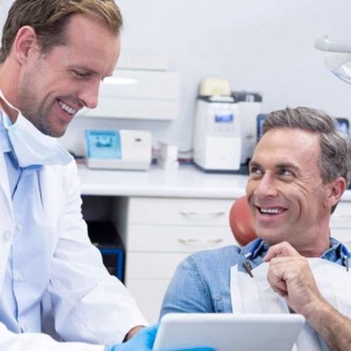 A dentist is talking to a patient in a dental chair while looking at a tablet.