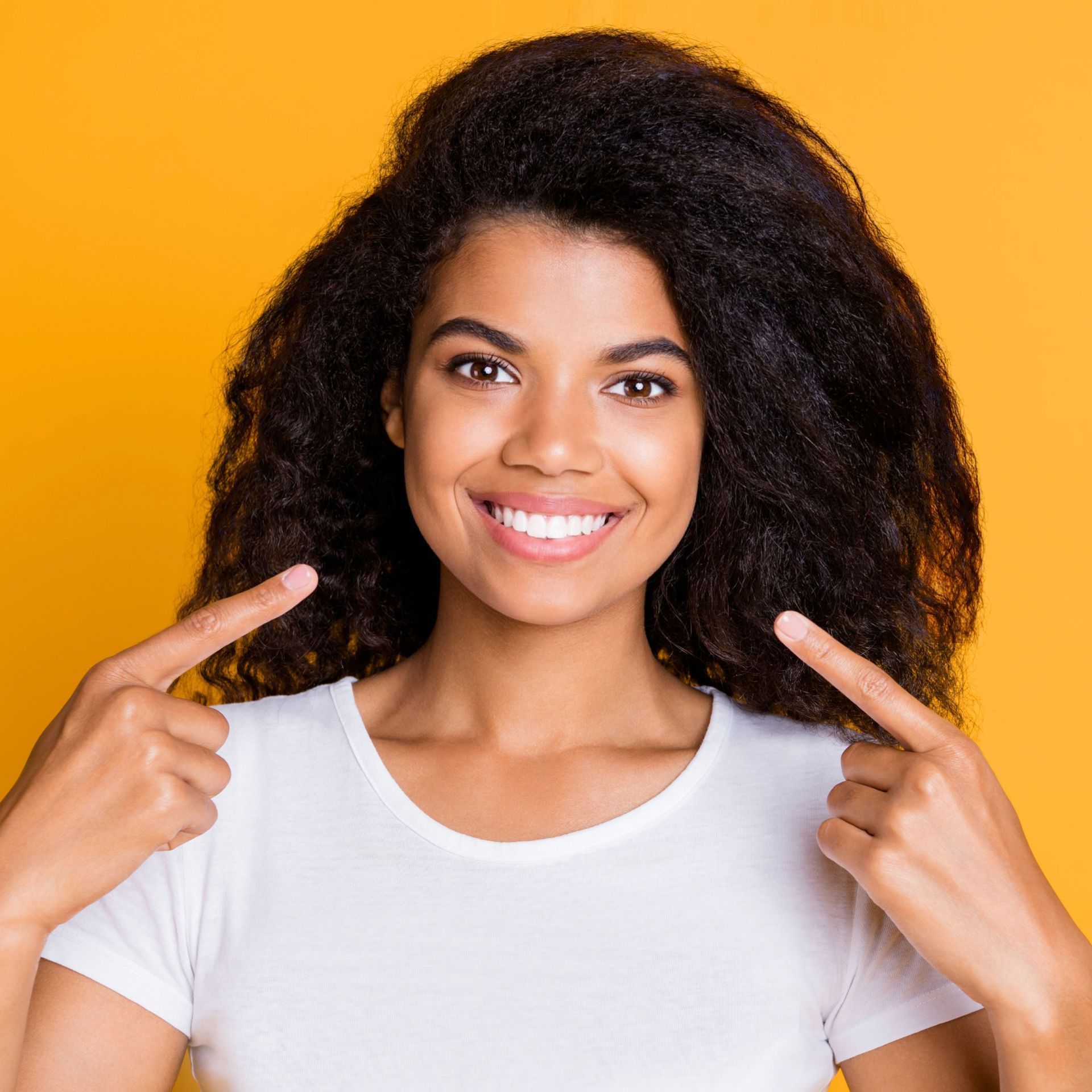 A woman with curly hair is smiling and pointing at herself.