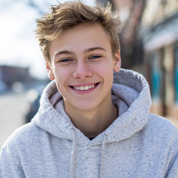 A smiling person with messy light hair wears a gray heathered hoodie in a blurred outdoor setting.