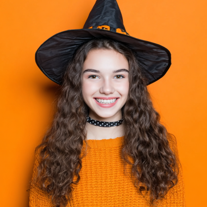 Girl wearing a witch hat and orange sweater smiles against an orange background.
