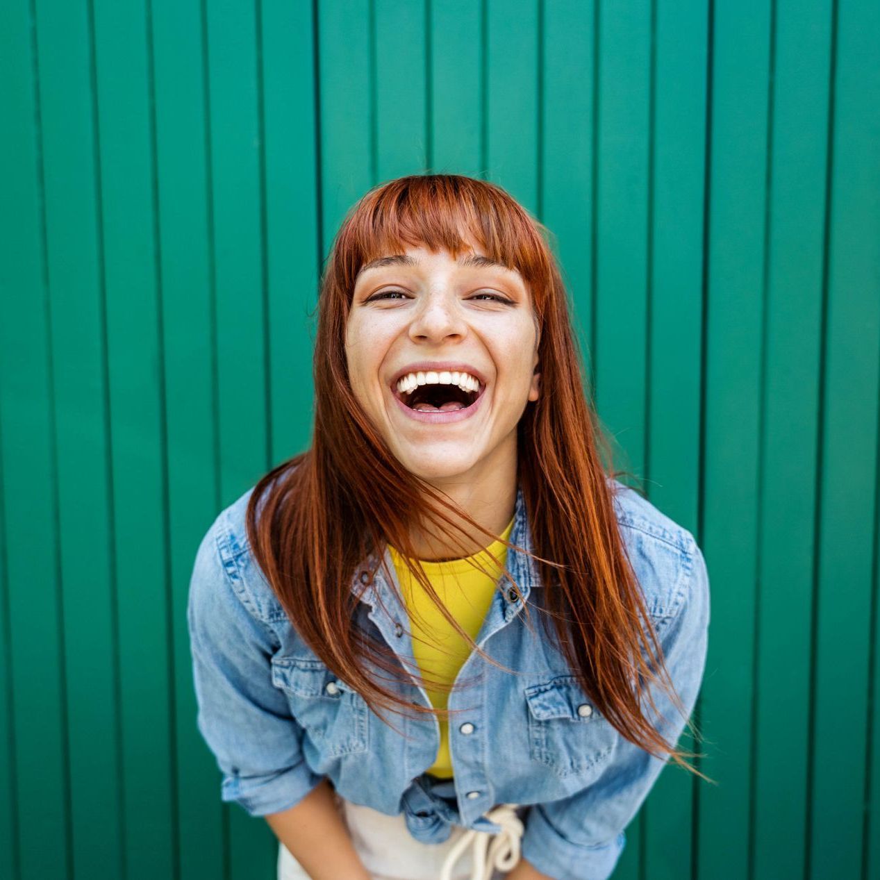 A woman with red hair is laughing in front of a green wall.