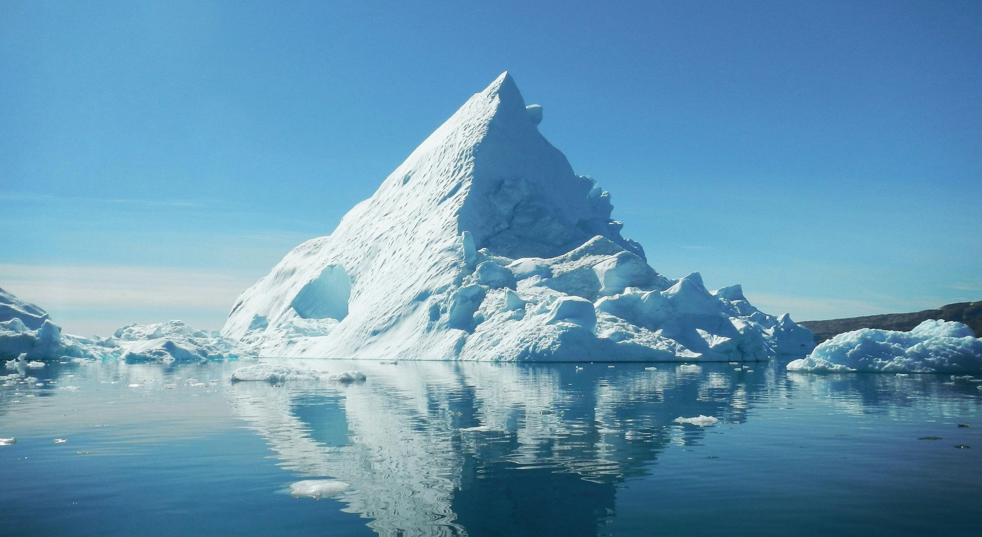 Iceberg in blue water, reflecting in the calm sea under a bright blue sky.