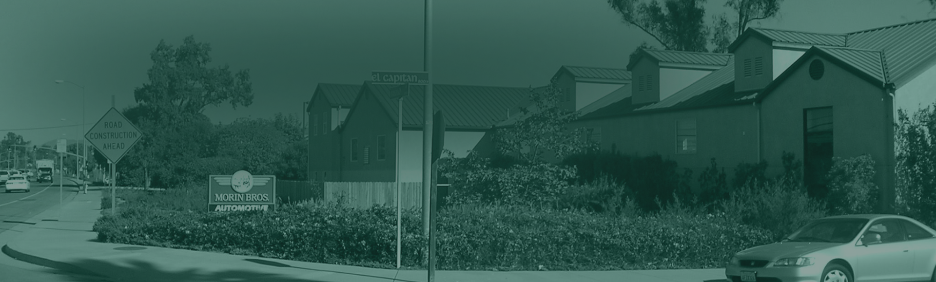 Roadside view with buildings, greenery, sign, and a car. Overcast and muted green tones. | Morin Brothers Automotive