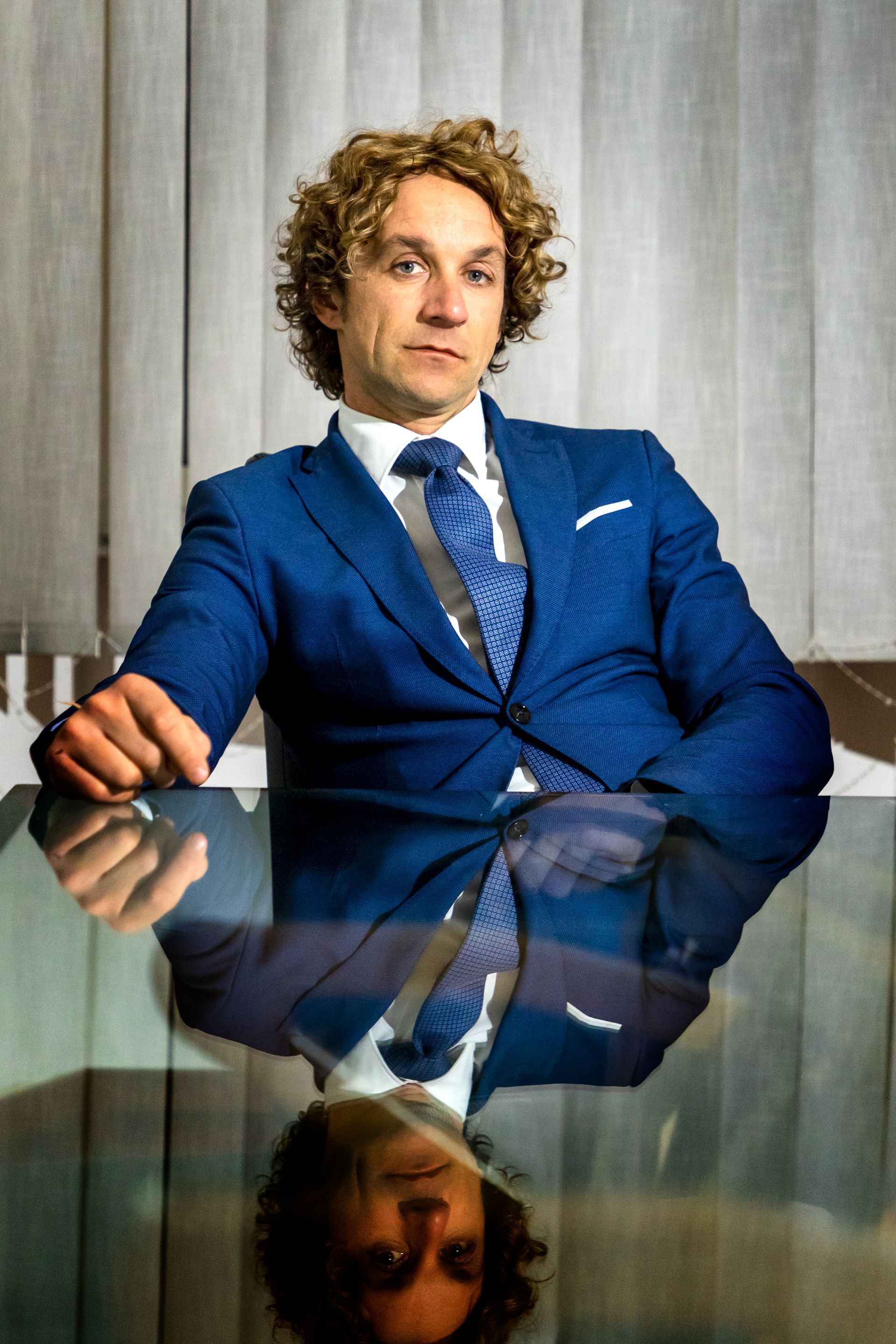 Un uomo in abito blu è seduto a un tavolo di vetro, riflesso sul piano del tavolo, sullo sfondo di una tenda neutra.