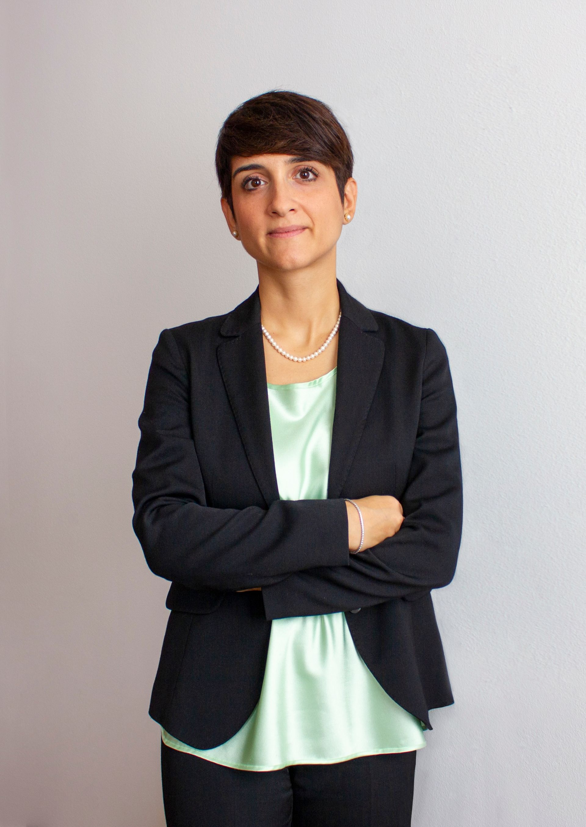 Persona in piedi con le braccia incrociate, che indossa un blazer nero sopra una maglietta color menta su sfondo grigio.