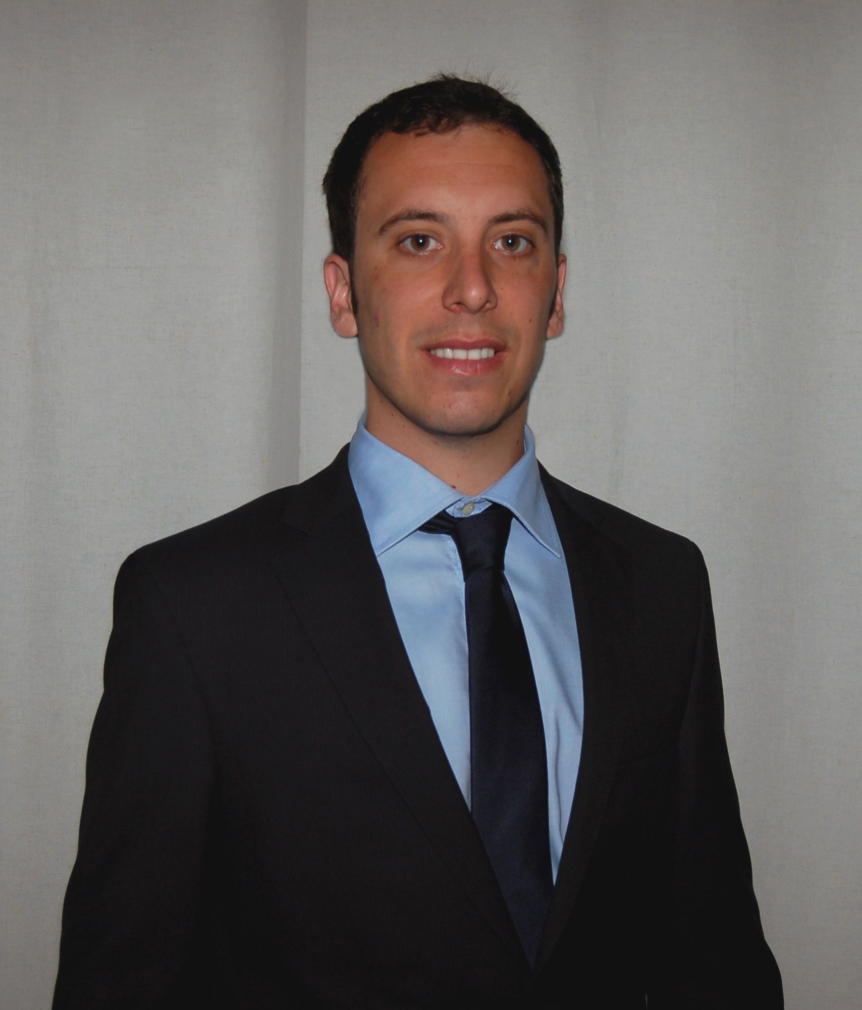 Uomo in abito nero e camicia blu in posa davanti a una tenda di luce.