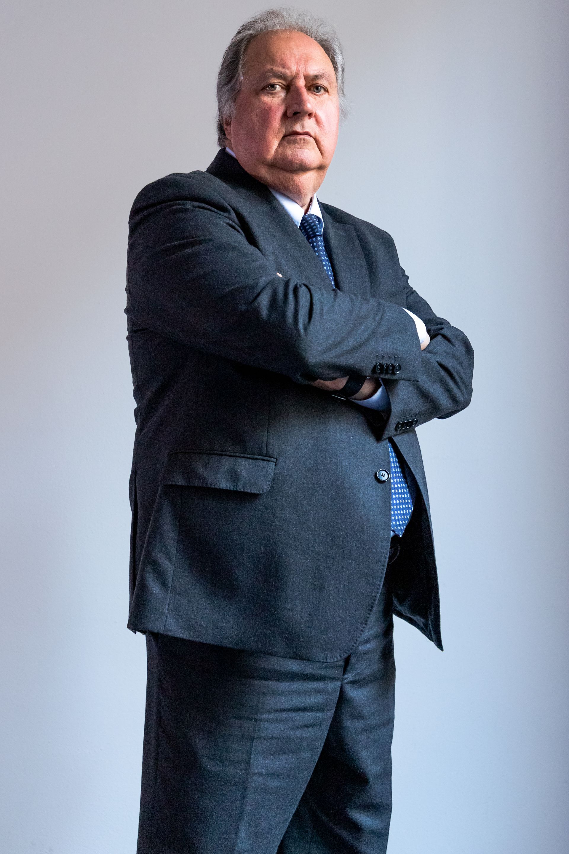 Uomo in abito scuro in piedi con le braccia incrociate contro un muro grigio uniforme.