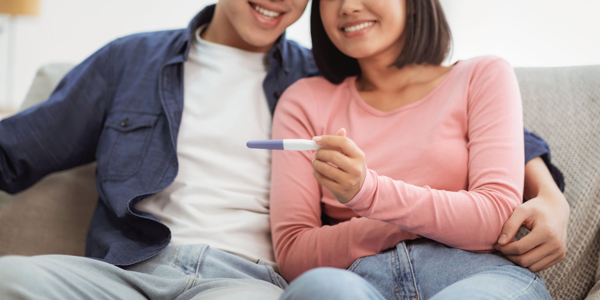 Couple smiling, holding a positive pregnancy test on a couch.