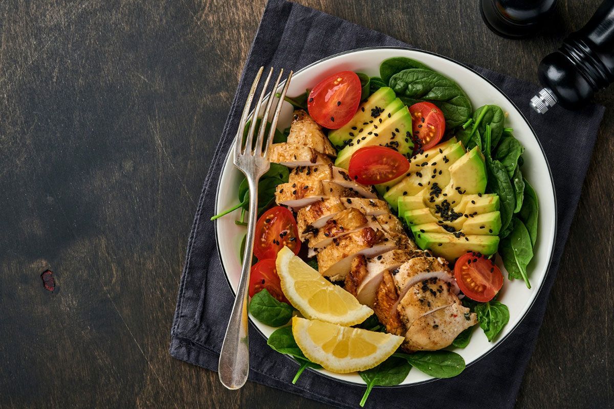A bowl of salad with chicken , avocado , tomatoes , spinach and lemon slices.