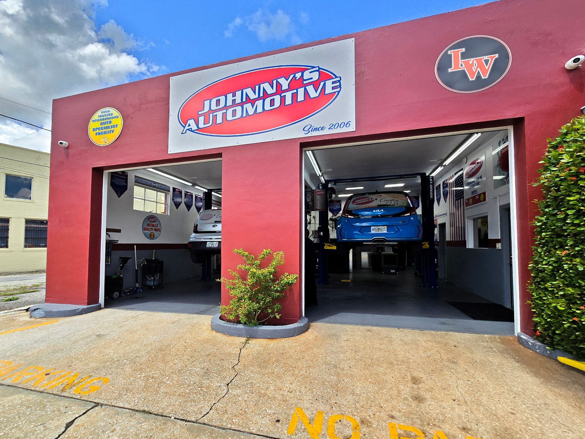 Johnny's Automotive shop with two garage bays. A blue car is on a lift. Red building, blue sky. | Johnny's Automotive