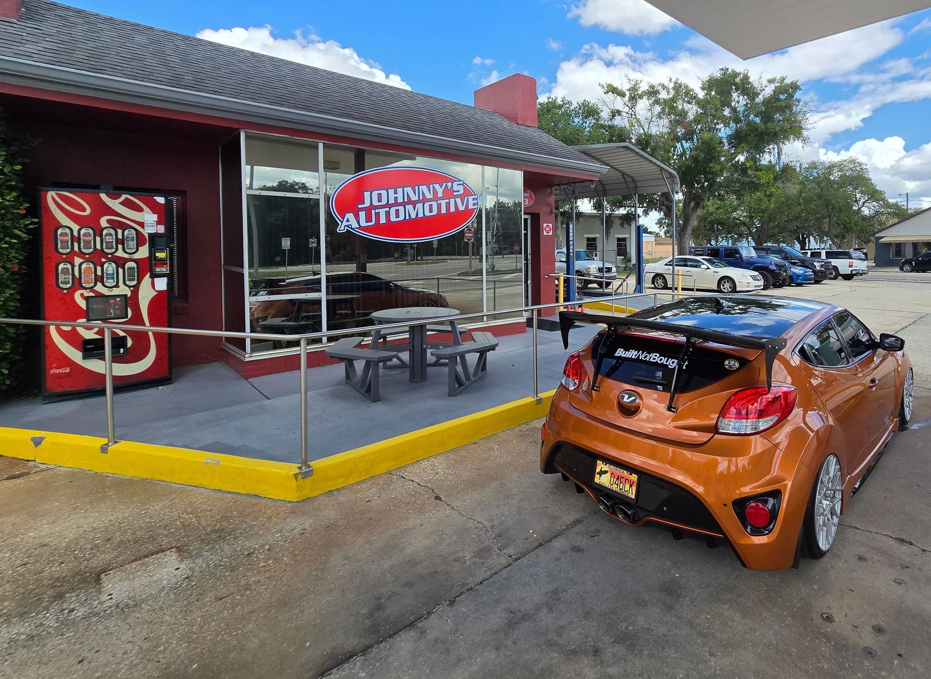 Orange car parked outside Johnny's Auto Repair. Red building, Coca-Cola machine, cloudy sky. | Johnny's Automotive