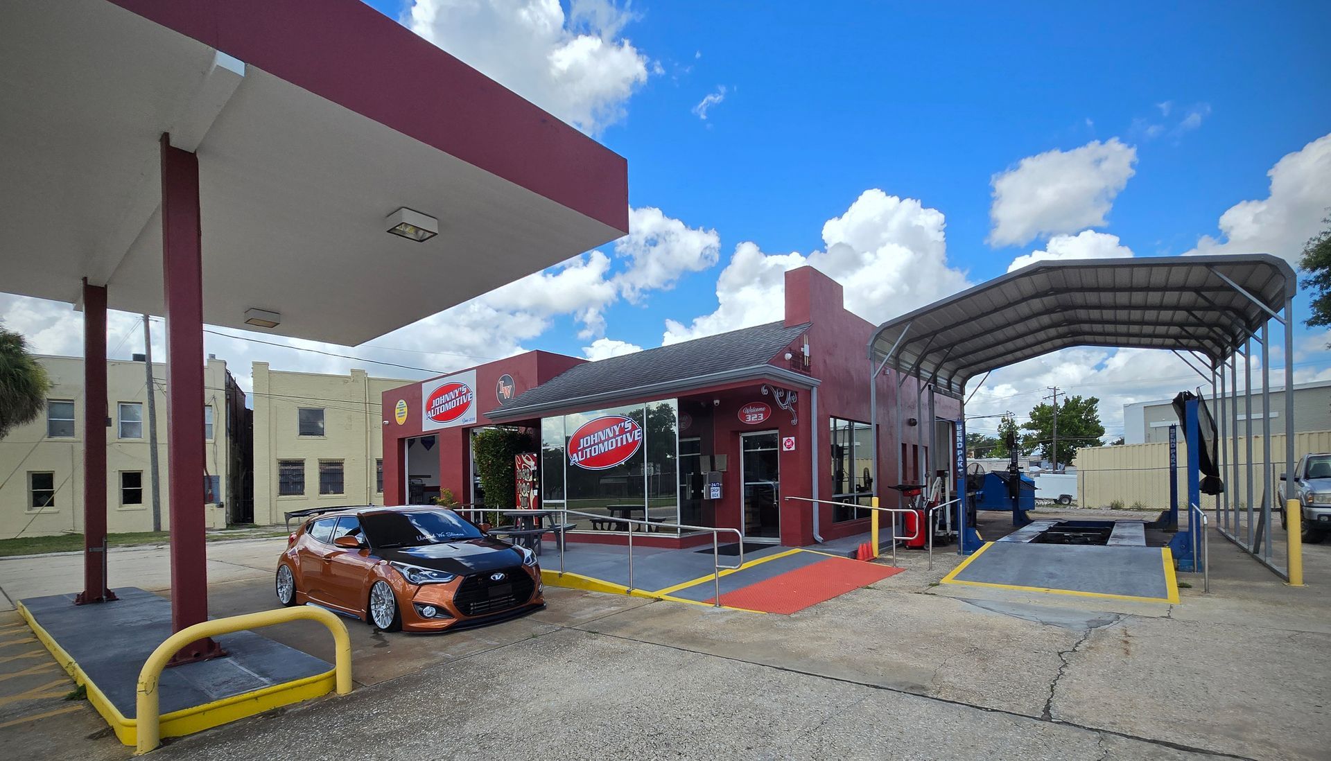 Red auto shop with gas pumps, a sports car, and a service bay under a blue sky. | Johnny's Automotive