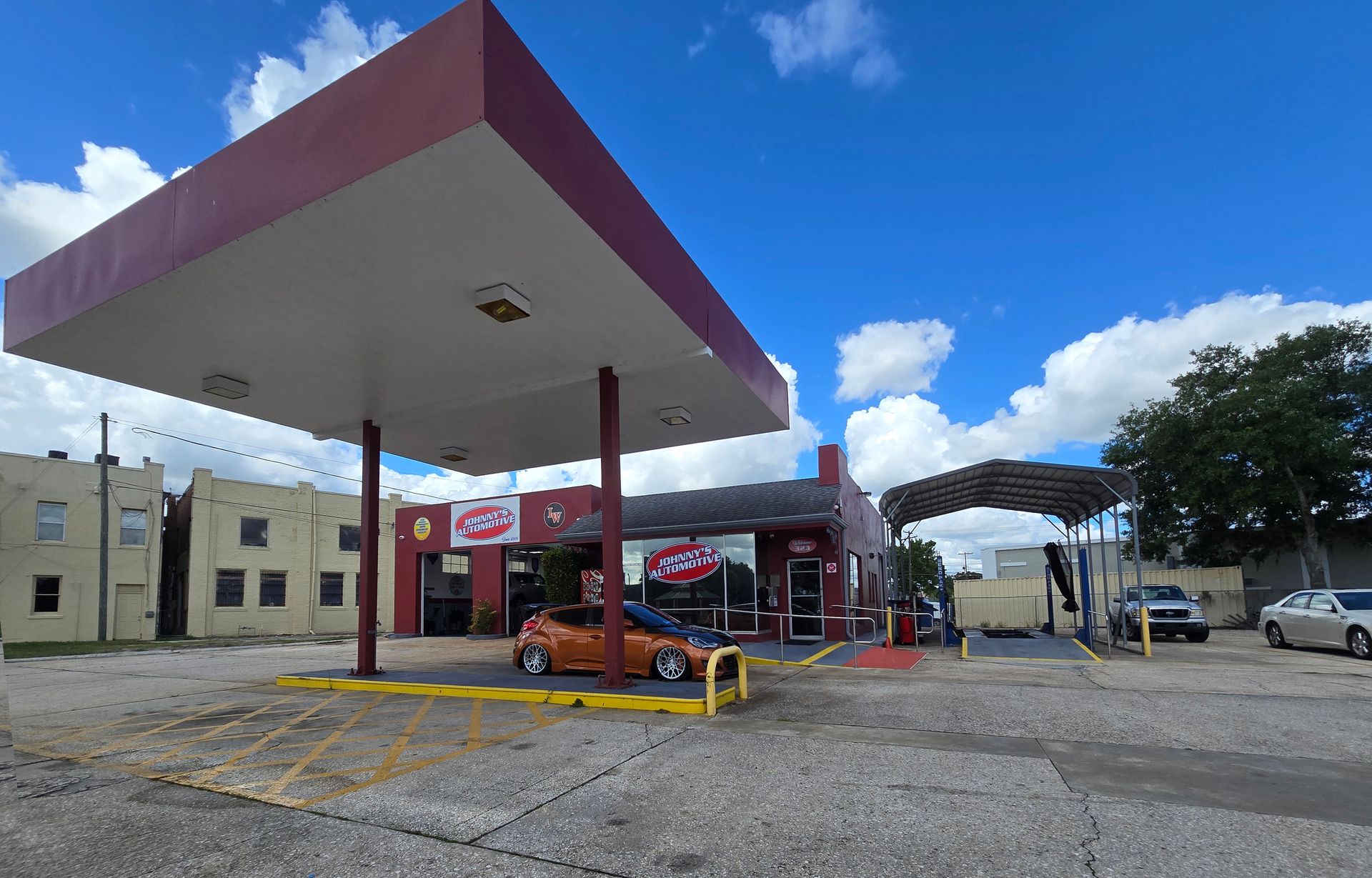 Red gas station with a car, blue sky, and surrounding buildings. | Johnny's Automotive