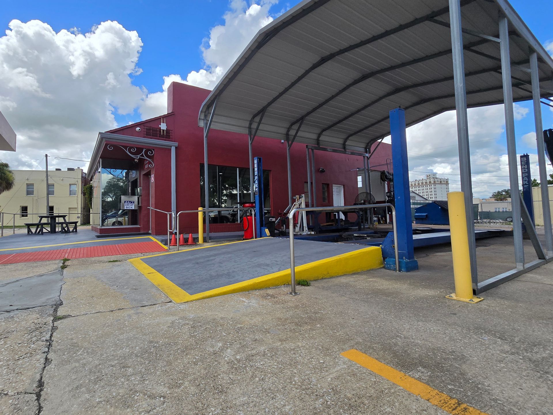 Red building with car lift under a canopy, yellow ramp. Outside, sunny day. | Johnny's Automotive