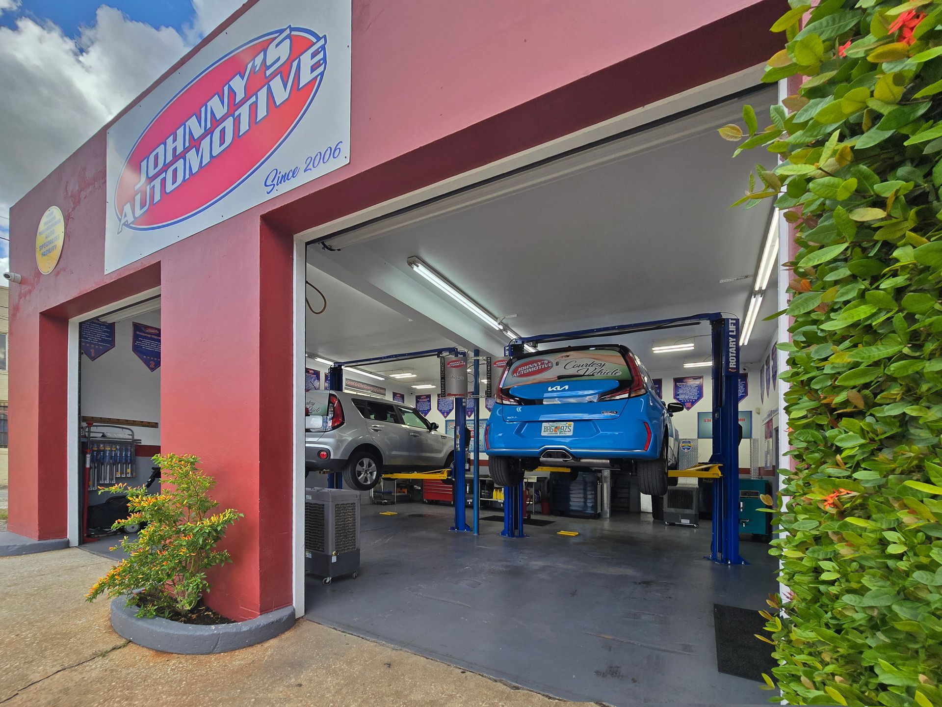 Jimmy's Automotive shop with a blue car on a lift and another vehicle inside. Red building with open bays. | Johnny's Automotive
