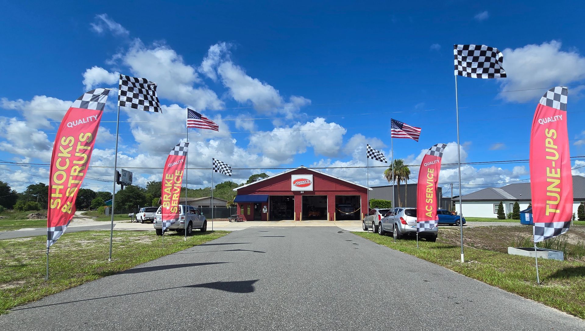 Auto repair shop with flags; red building, blue sky, driveway, and cars. | Johnny's Automotive